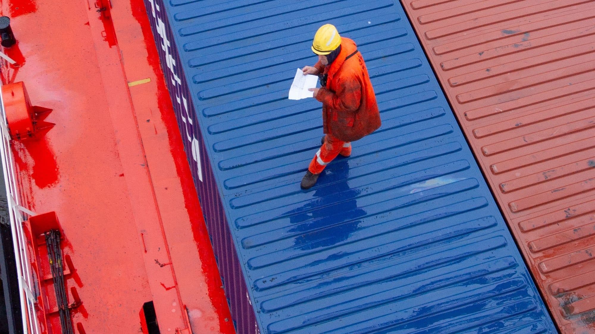 Ein Arbeiter in orangem Arbeitsanzug und gelbem Schutzhelm steht auf einem blauen Container an Deck eines Schiffes und prüft ein Blatt mit Unterlagen
