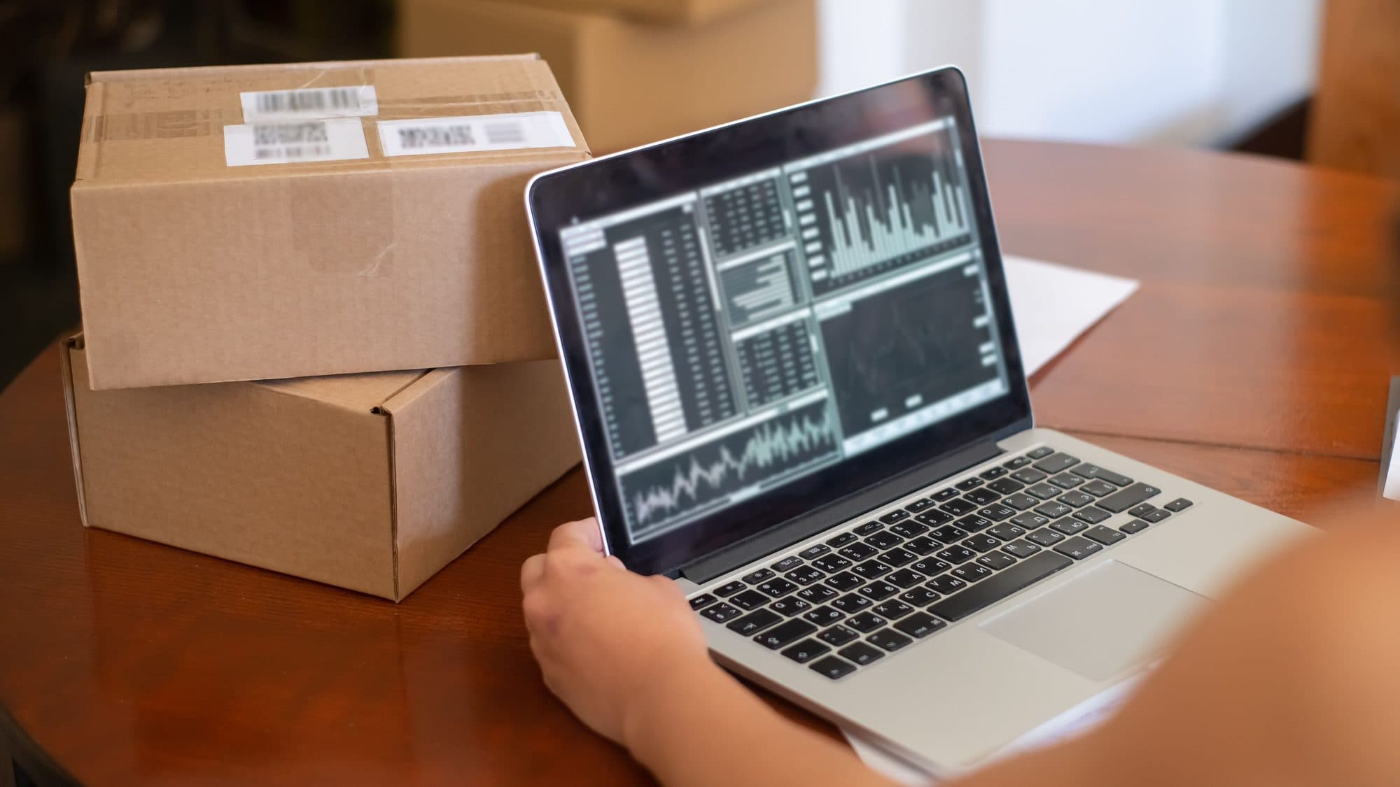 A hand rests on a laptop showing tables of figures and bar charts, next to two labelled shipping parcels on a wooden table