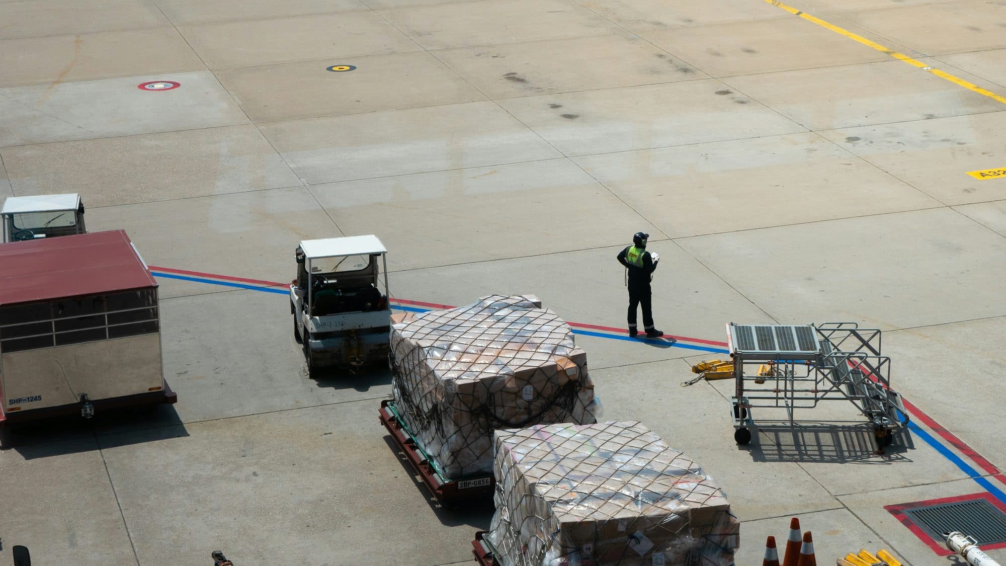 Zwei mit Netzen gesicherte Luftfrachtpaletten auf Dollys auf dem Vorfeld eines Flughafens, daneben ein Bodenabfertiger in Warnweste, ein Schlepper, eine Ladeplattform und ein Luftfrachtcontainer
