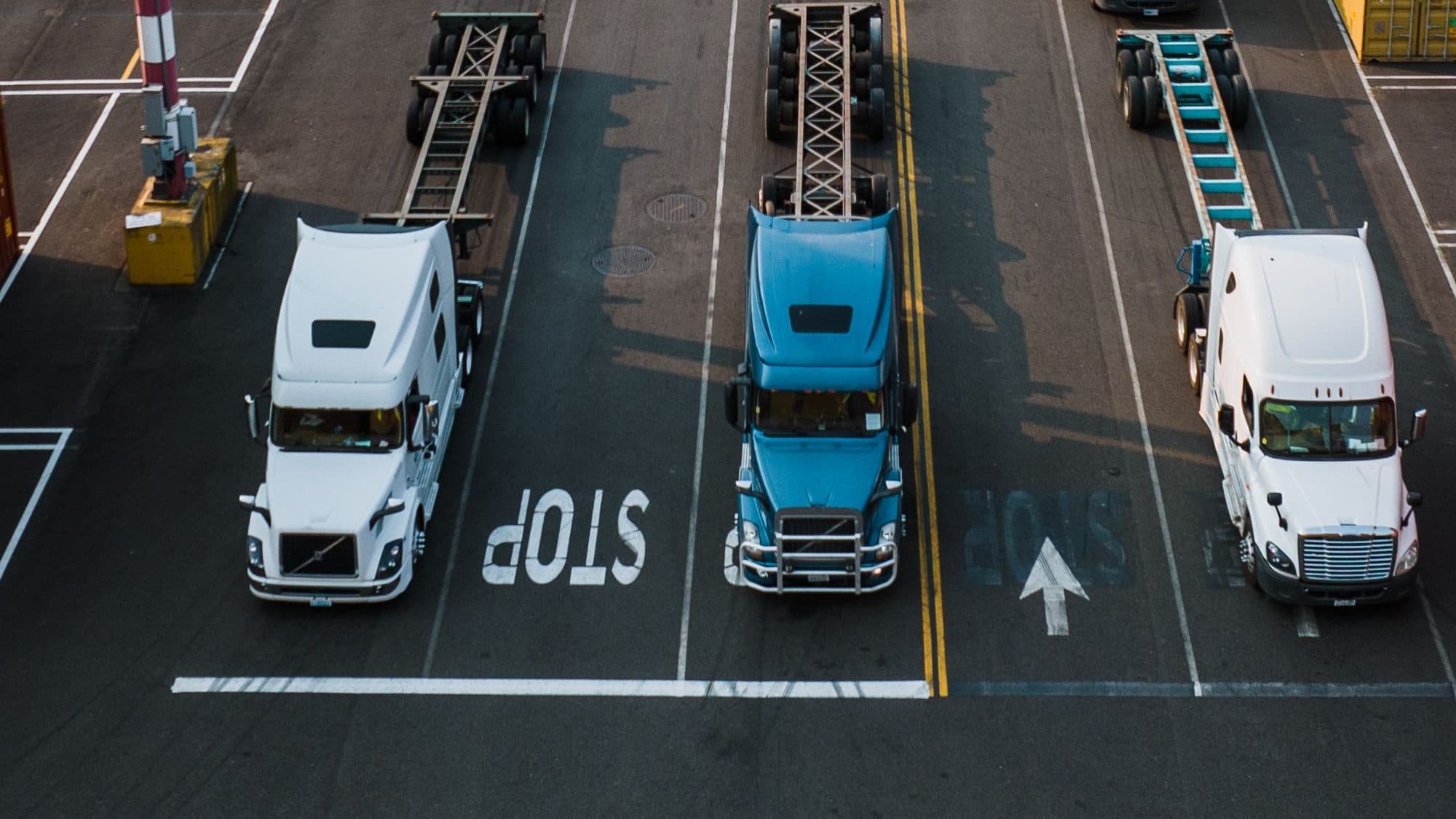 Three tractor units pulling empty container chassis wait at the stop line of their lanes at a port container terminal, with more trucks and chassis queuing behind them