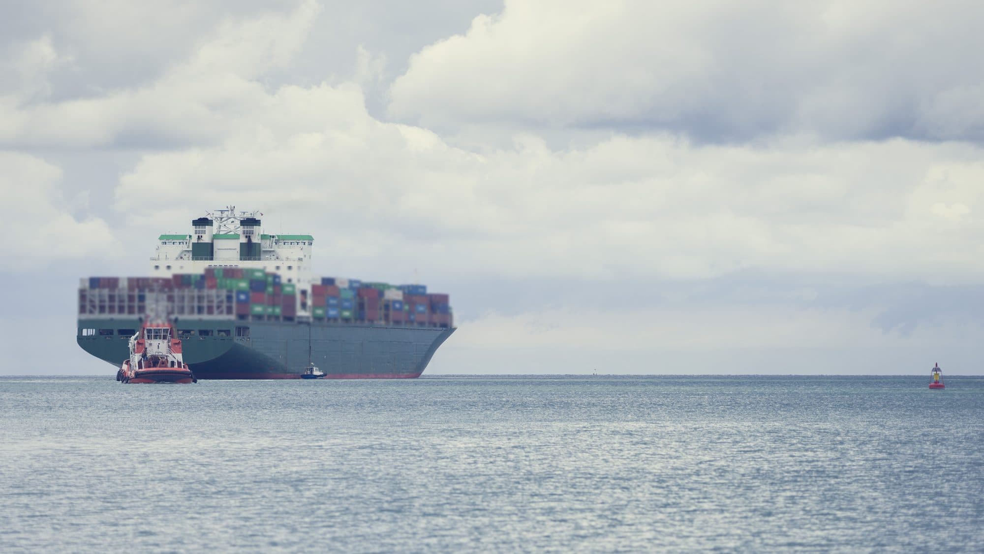 A loaded container ship under way on grey-blue water under a cloudy sky, seen from astern, with a red tug at its stern and a small pilot boat alongside