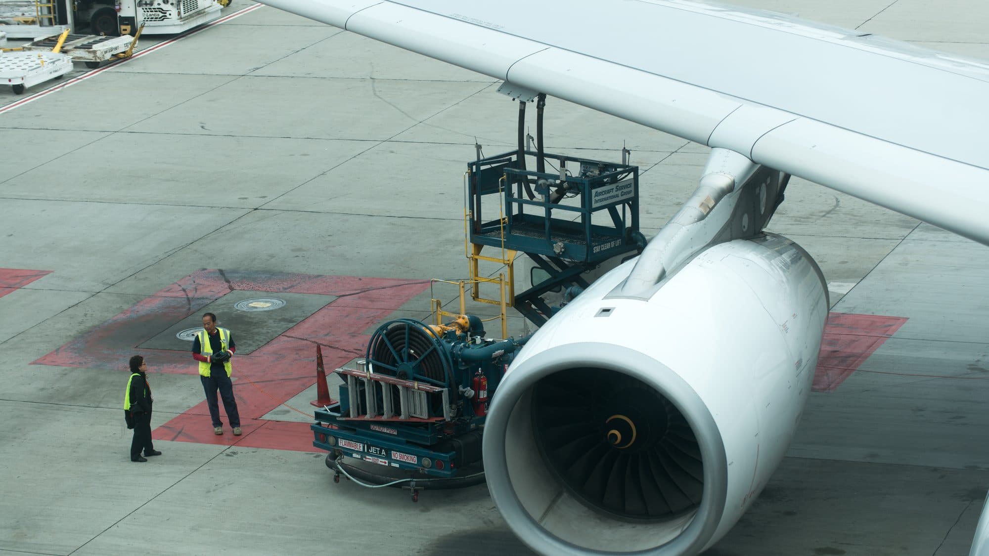 Two ground staff in high-visibility vests stand on the apron beside a hydrant dispenser refuelling an airliner under its wing
