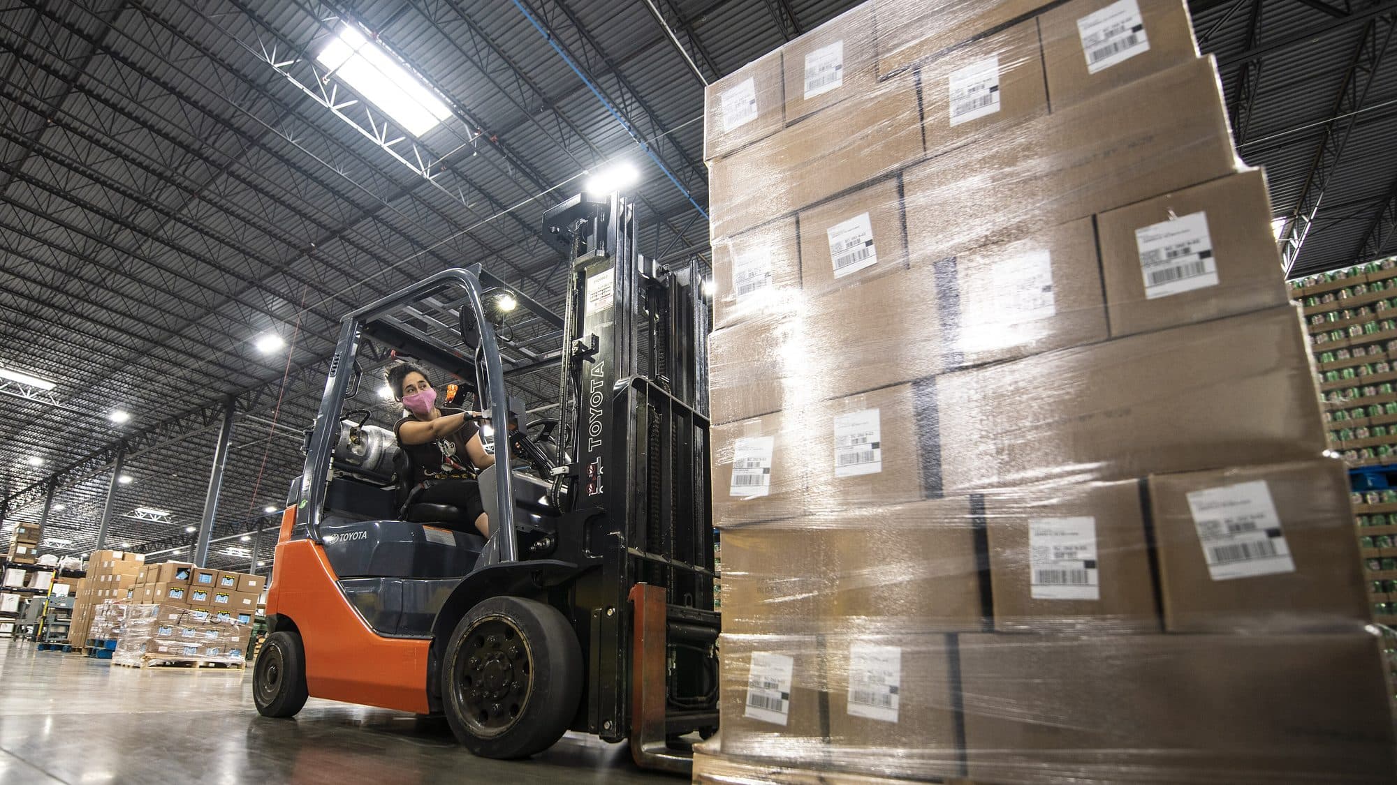 A forklift driver moving a pallet of labelled, shrink-wrapped boxes through a warehouse