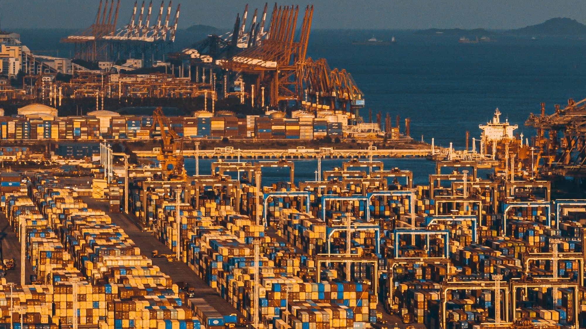 Elevated view at golden hour over a container terminal storage yard in Xiamen, with long rows of stacked containers, rubber-tyred gantry cranes and quay cranes along the waterfront